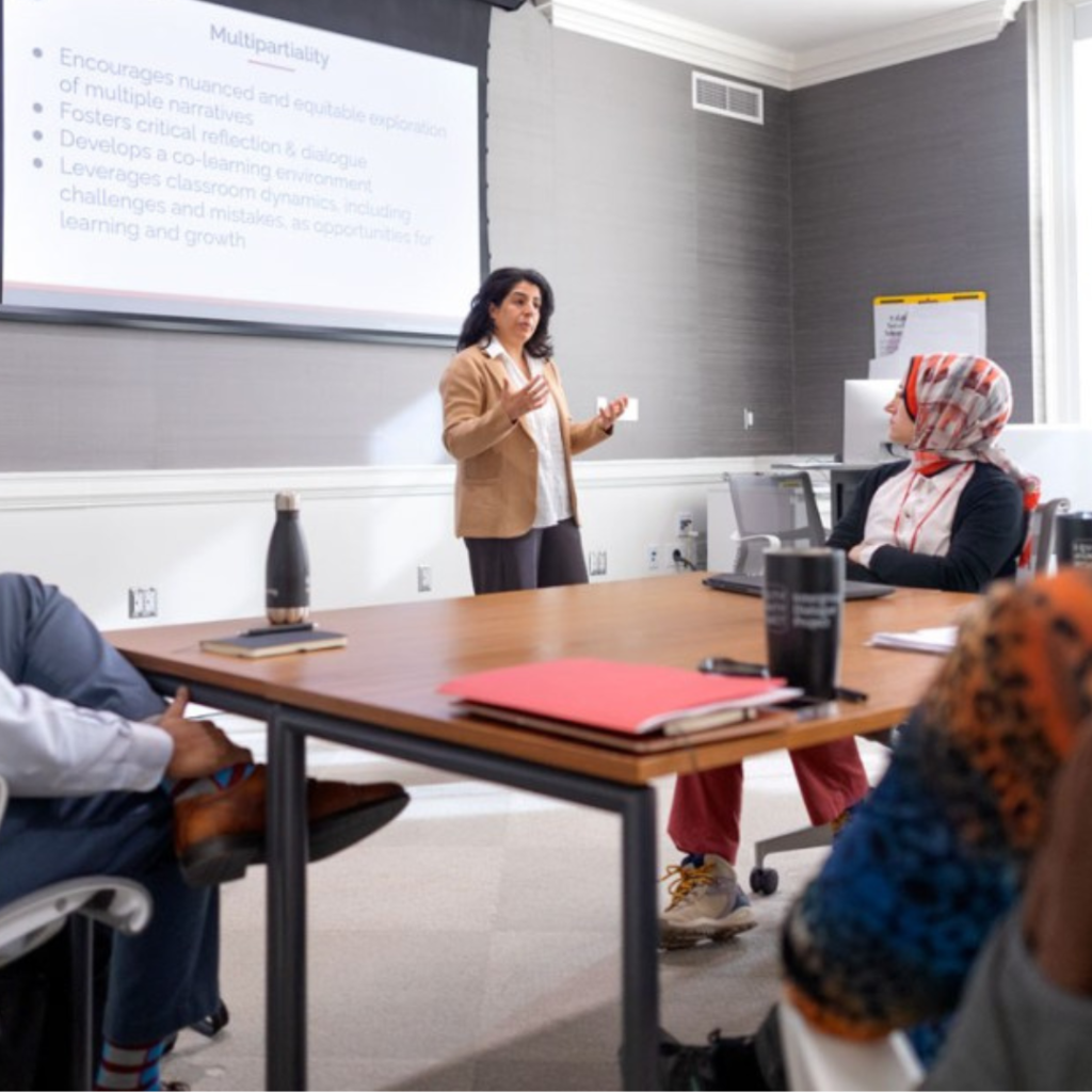 A woman lectures in front of a projector screen depicting several bullet points under the word "maltipartiality." Three people listen at a table, including a woman wearing a head-covering.