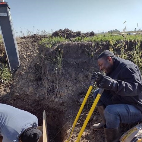 IIE-SRF fellow Eric Zama holds two shovels, sitting in a trench dug into the earth as part of his research.